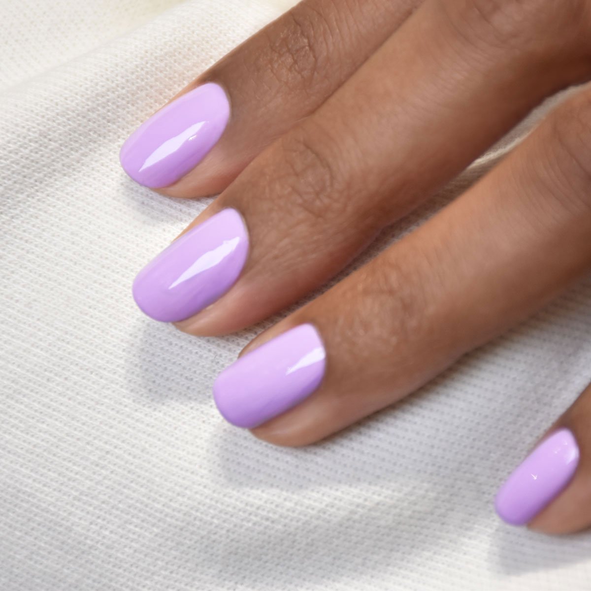 A close-up of a hand with neatly manicured nails painted in glossy lilac Lisa Lilas polish by Manucurist, resting on white textured fabric.