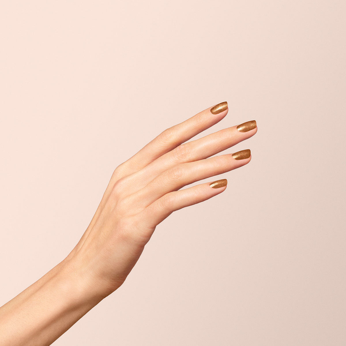 A hand with slender fingers is raised against a neutral background, showing nails painted with Bronzé, a shimmering copper nail polish from Manucurist.