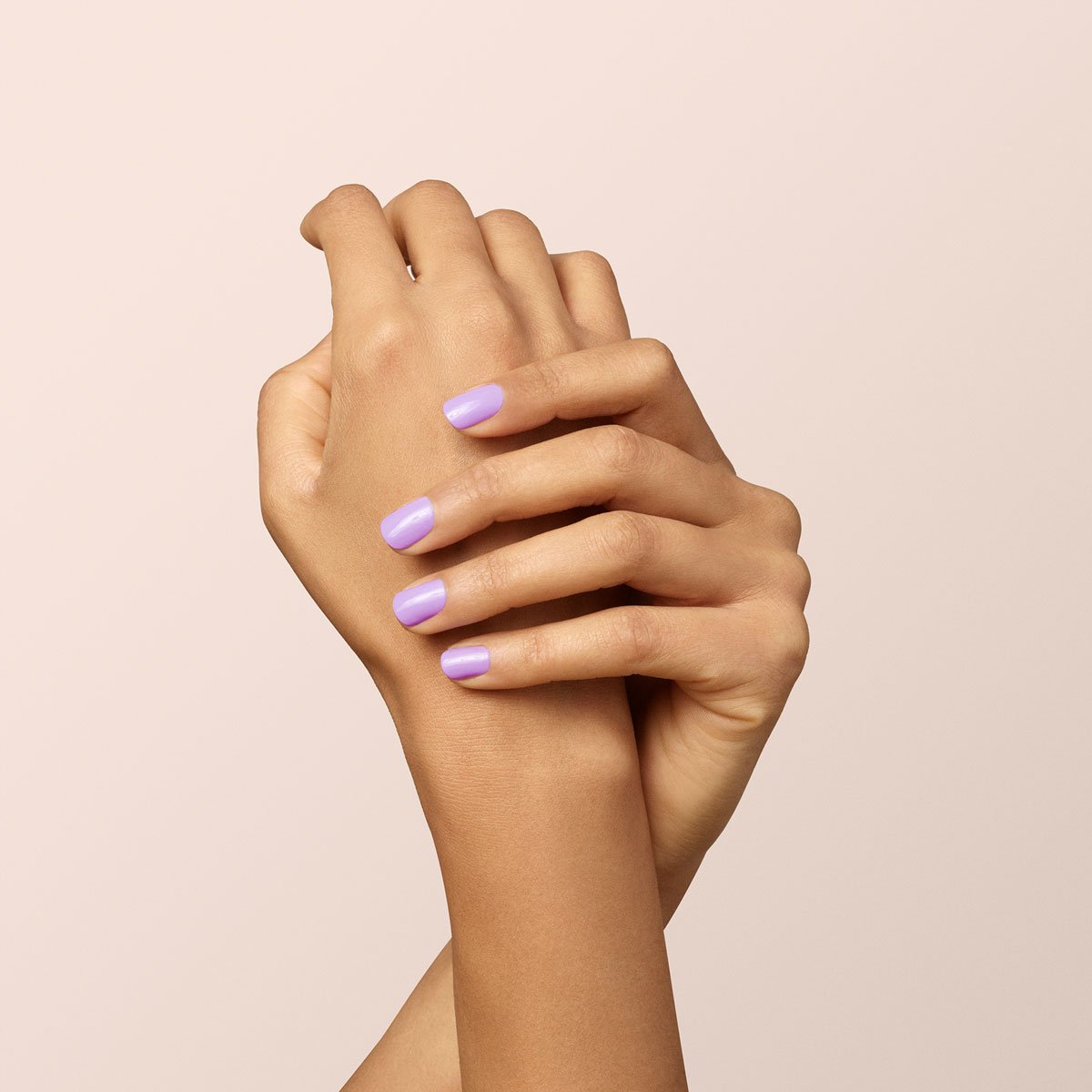 Two hands with light brown skin are gently clasped together, displaying neatly manicured nails painted in Manucurist’s Lisa Lilas lilac nail polish against a neutral beige background.