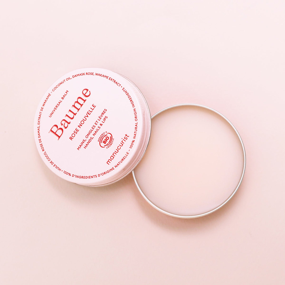 Un pot rond et ouvert de Baume Nourrissant Naturel Manucurist - Cosmos Organic repose sur une surface rose clair. Le couvercle au marquage rouge est posé à côté, révélant le baume lisse à l’intérieur — parfait pour réparer les lèvres et prendre soin naturellement de la peau.