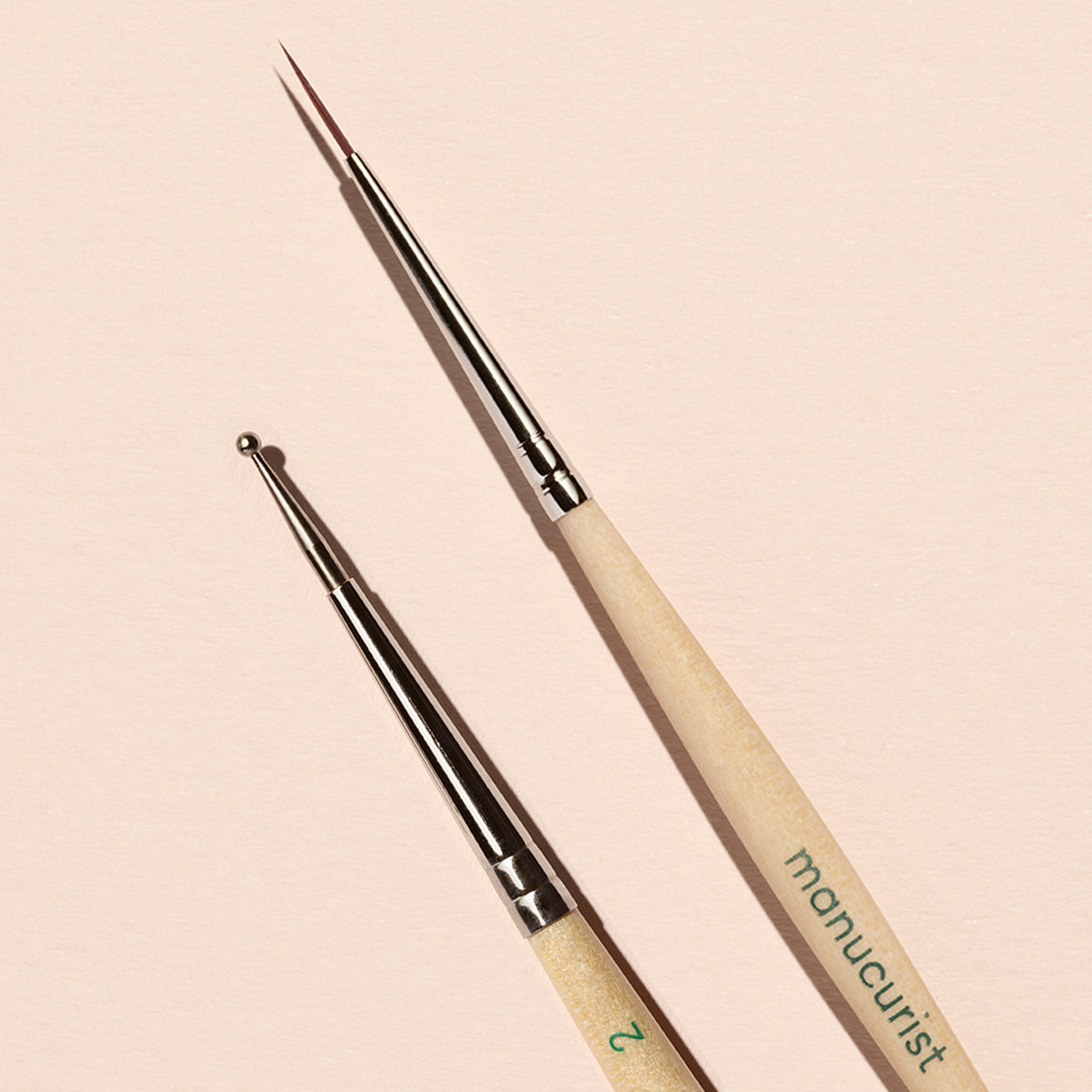 Le duo Dotting Tool Nail Art Liner Brush de Manucurist présente des manches en bois clair et des embouts argentés, disposés sur un fond rose pâle.