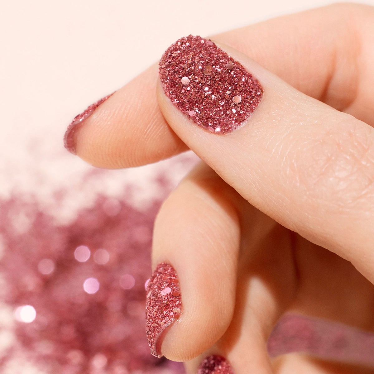 Un gros plan d’une main aux ongles ornés de Pink Quartz Biodegradable Glitter de Manucurist, d’un superbe Nail Art et d’accents scintillants dispersés en arrière-plan.