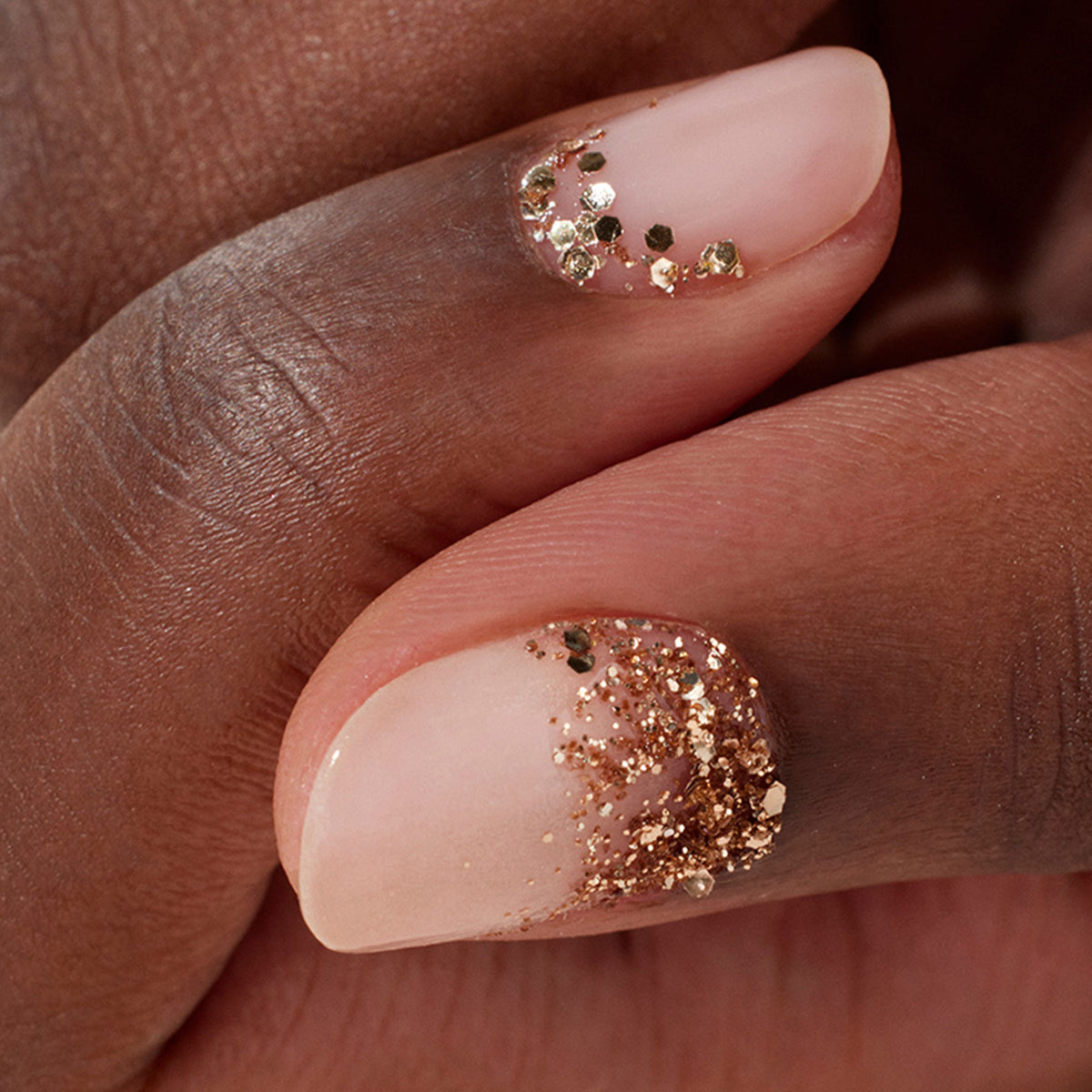 Two fingers with nude nails feature eco-friendly Manucurist Rose Gold Biodegradable Glitter accents on the cuticles, creating a chic and elegant nail art look.