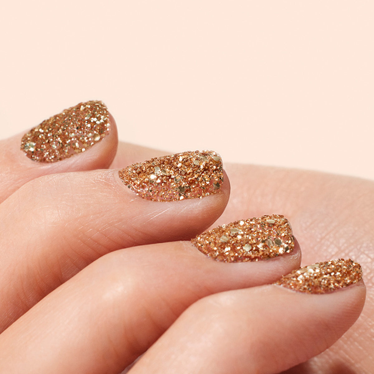 Close-up of a hand with nails painted in Manucurist Rose Gold Biodegradable Glitter polish, set against a soft, neutral background.
