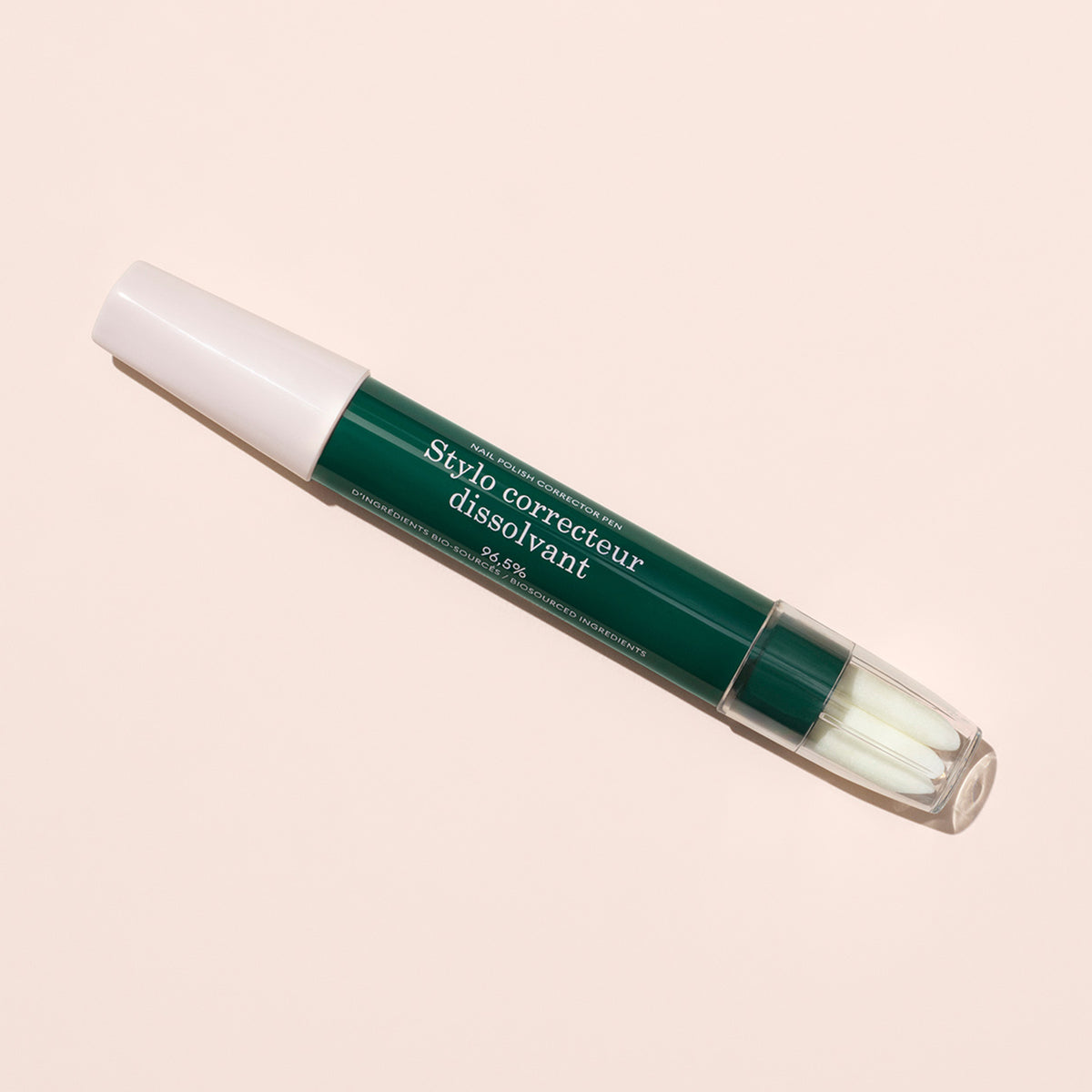 A Manucurist-test + Corrector Pen, green and white with "Stylo correcteur dissolvant" on it, lies diagonally on a light beige background. The cap covers one end, while a clear cover reveals its fine tip for precise gel polish correction.