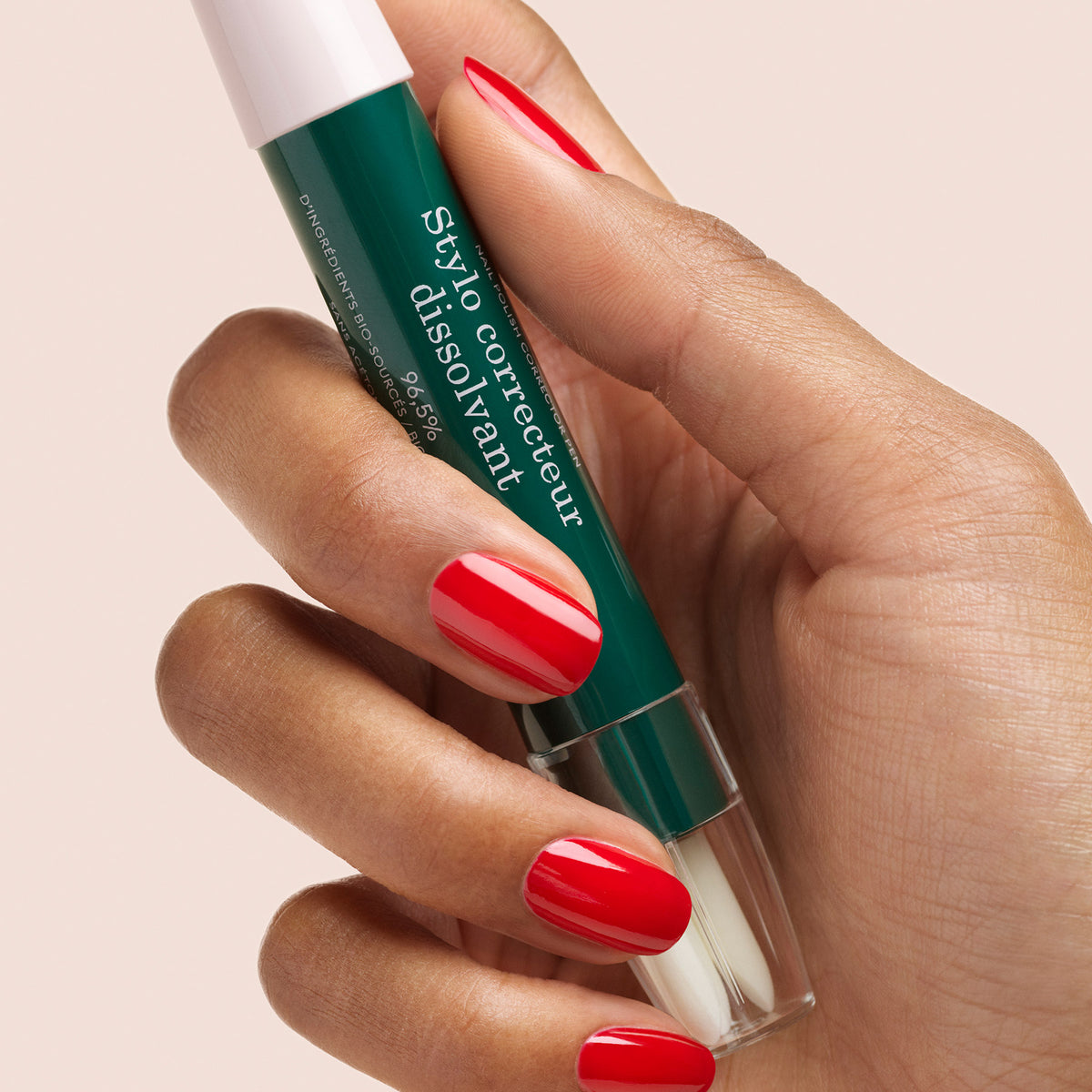 Une main aux ongles rouges impeccablement manucurés tient le Manucurist-test + Corrector Pen, un stylo correcteur de vernis à ongles vert, sur un fond beige clair.