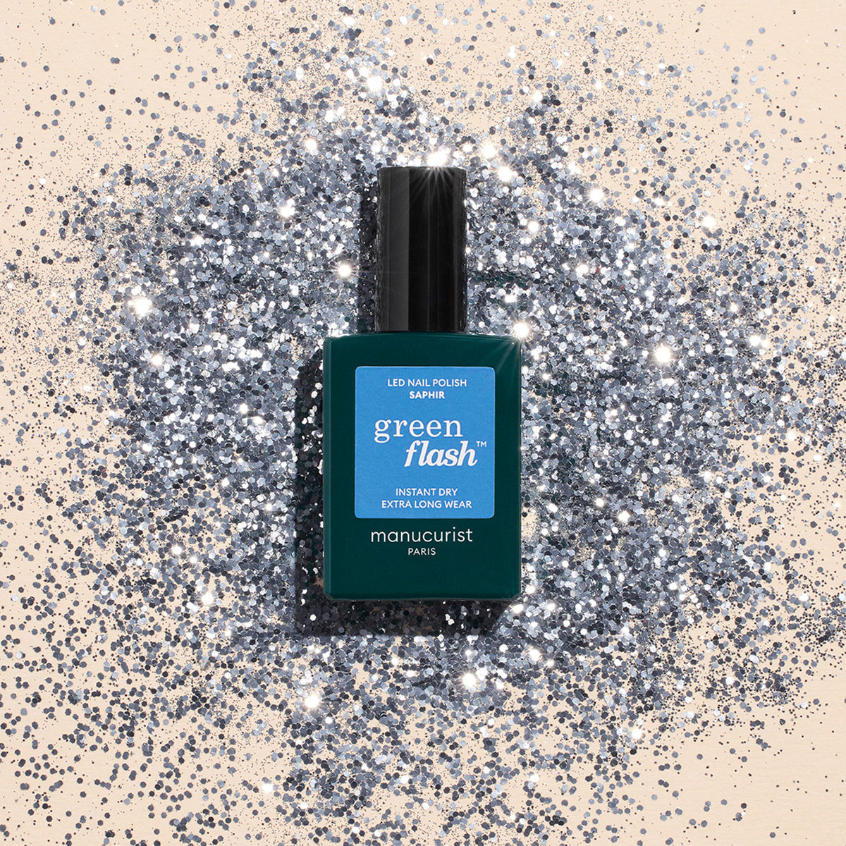 A teal bottle of Manucurist Saphir gel polish rests on a surface dusted with shiny silver glitter, creating a sparkling, glamorous look.