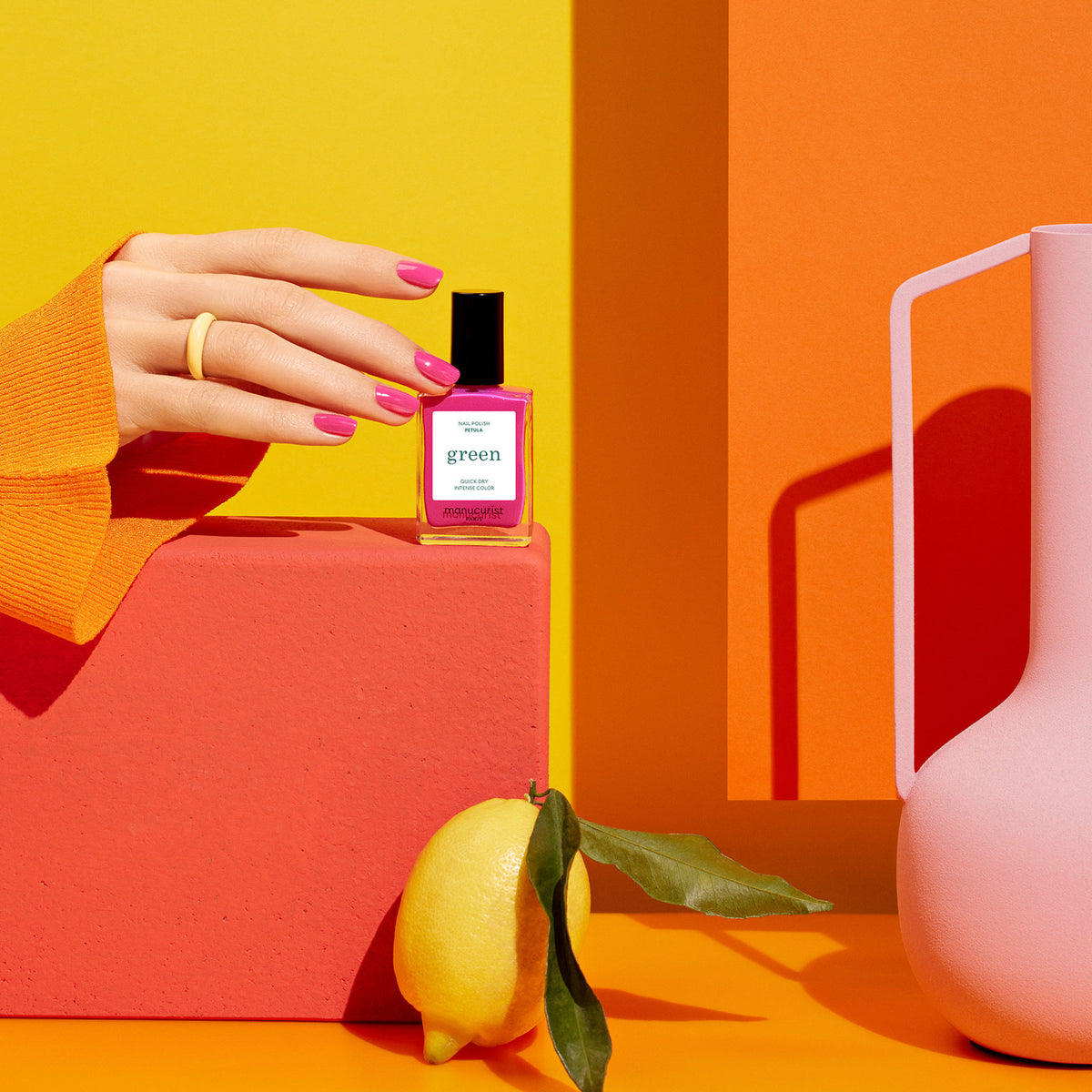 A hand with pink nails holds a pink Pétula perfume bottle by Manucurist against a vibrant orange-yellow background, beside a pink vase, lemon, and coral block. Blue undertones add depth to the lively scene.
