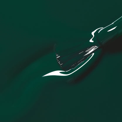 A close-up of a paintbrush spreading Manucurist’s Emerald nail polish, a glossy deep forest green shade, with light reflecting off the wet surface.
