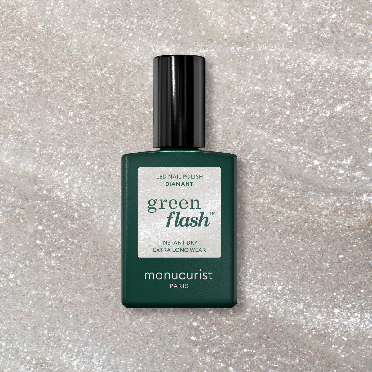 A bottle of Manucurist Diamant gel polish rests on a sparkly, light surface. The dark green bottle with a black cap and white label showcases the shimmering silver nail color.