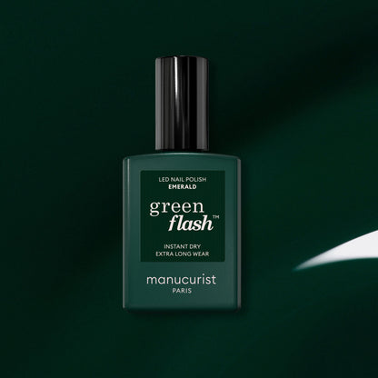 A bottle of Manucurist’s Green Flash LED gel nail polish in the shade Emerald sits centered on a dark green background. The label features the phrases “Instant Dry” and “Extra Long Wear,” emphasizing the gemstone-inspired polish’s rich, sophisticated appearance.