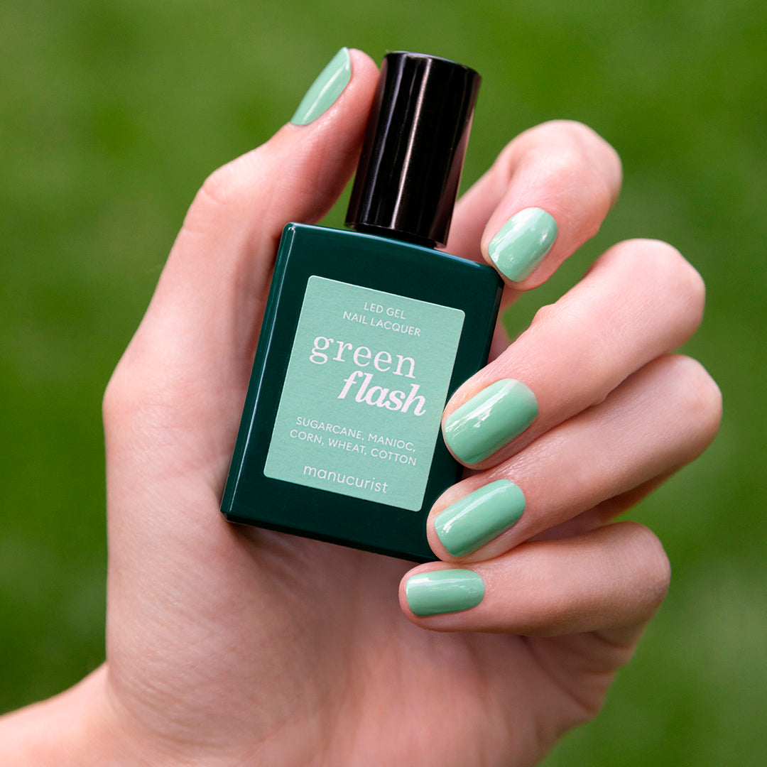 A hand with neatly manicured, mint green gel polish nails holds a bottle of Manucurist Amande LED gel nail lacquer against a blurred green background.