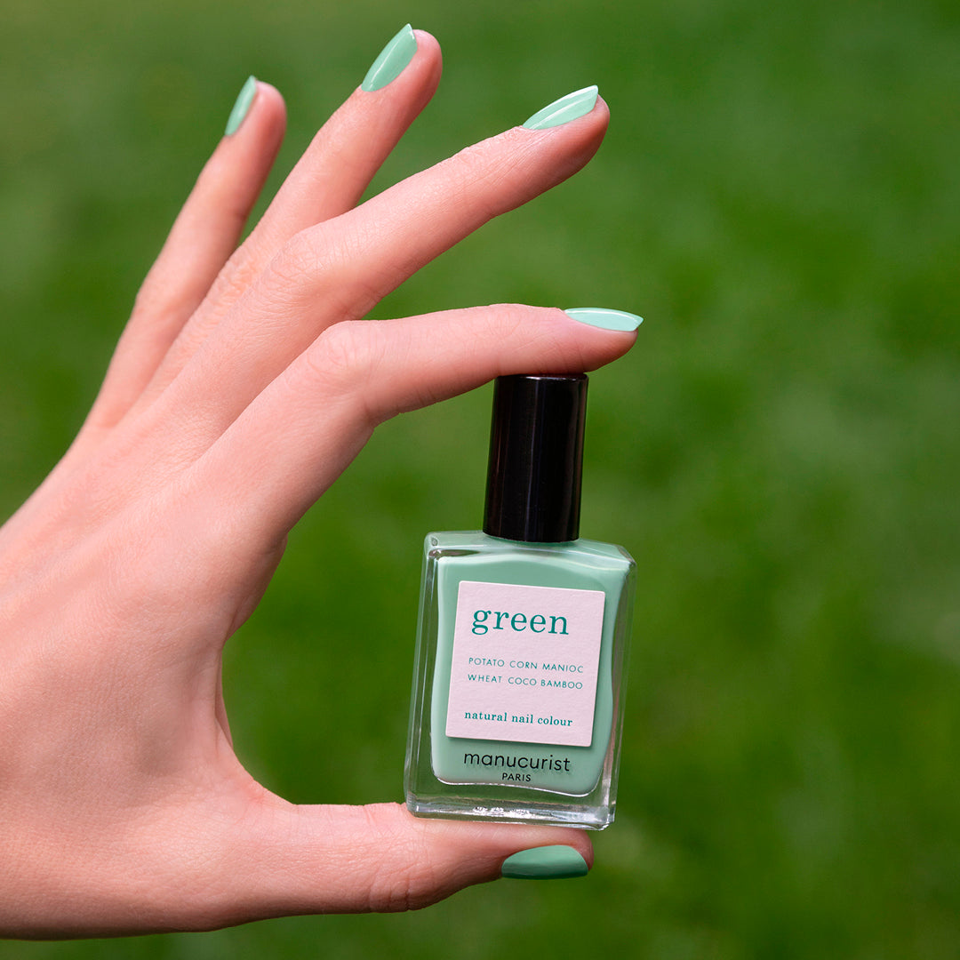Une main aux ongles vernis vert menthe tient un flacon de vernis Amande de Manucurist sur fond vert flou.