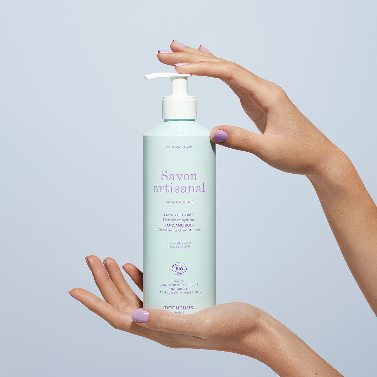 Une personne aux ongles peints en lavande tient un flacon pompe bleu clair du Organic Lavender Hand  Body Wash - Cosmos Organic de Manucurist contre un fond bleu clair, évoquant un parfum de lavande apaisant.