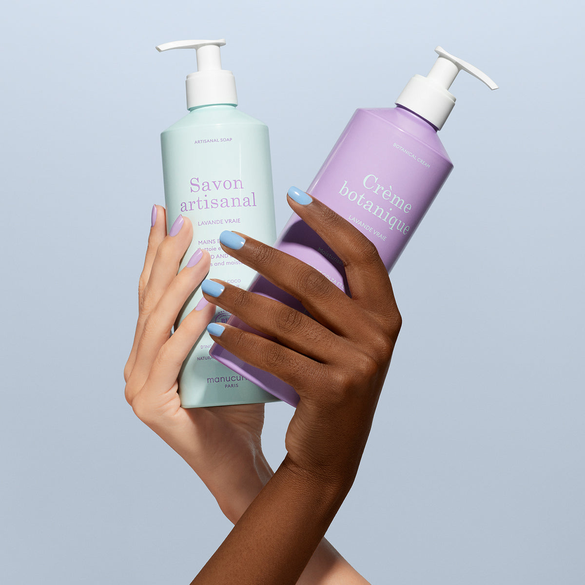Deux mains, l’une claire et l’autre foncée, avec des ongles bleu pastel assortis, tiennent le Duo Crème Botanique &amp; Savon Artisanal Lavande - Cosmos Organic de Manucurist sur un fond bleu clair.
