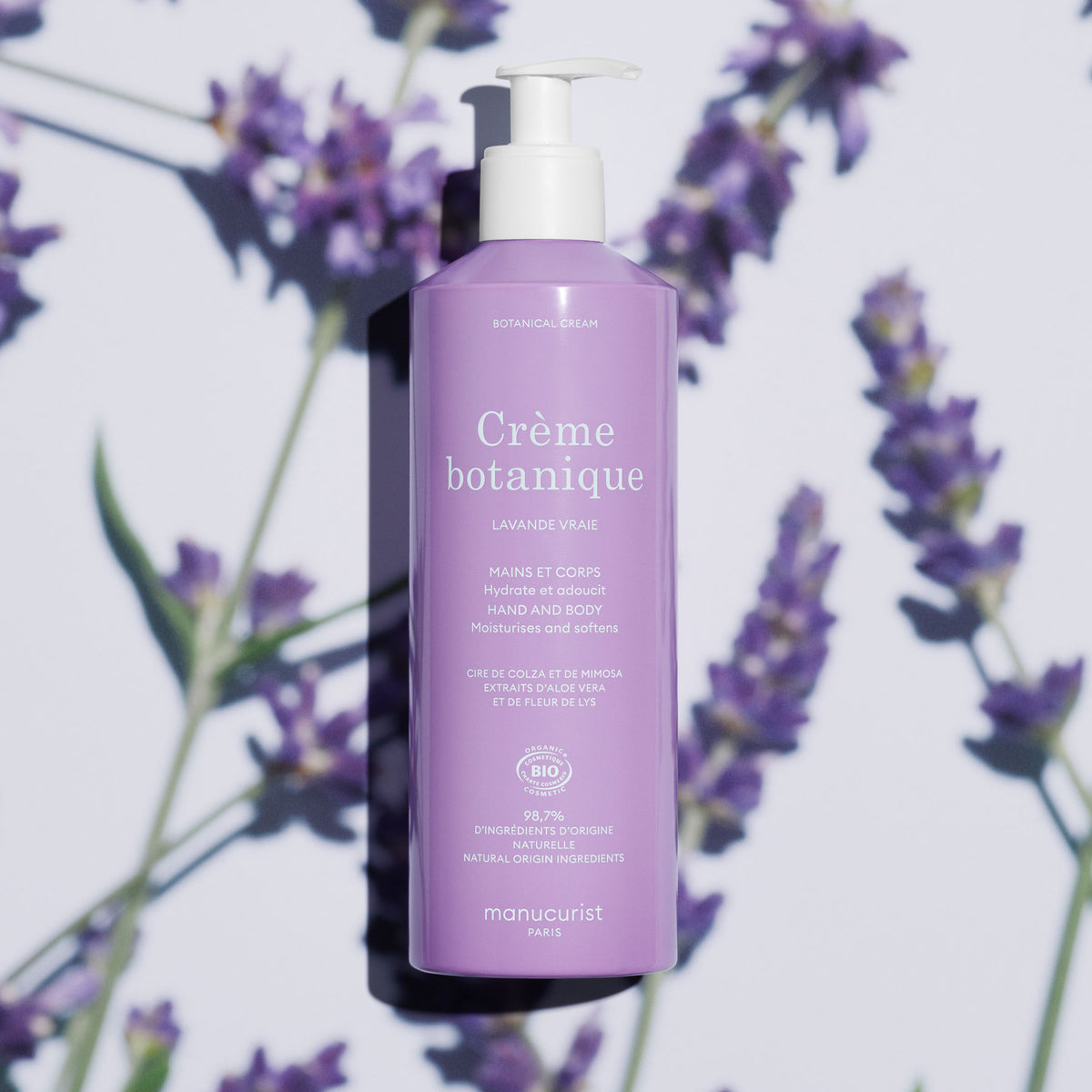Un flacon violet de crème pour les mains et le corps Soothing Lavender de Manucurist - Cosmos Organic, avec une pompe blanche, est posé sur une surface blanche, encadré de fleurs de lavande et d’ombres. L’étiquette française met en avant son essence botanique et sa formule hydratante.