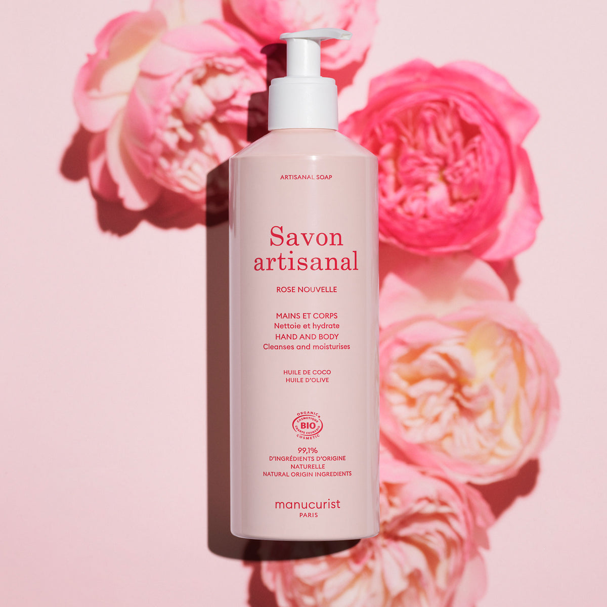 Un flacon rose de Manucurist Organic Rose Hand Body Wash - Cosmos Organic repose sur un fond rose avec des roses, son étiquette indiquant un délicat parfum de rose et des ingrédients naturels pour un soin luxueux des mains et du corps.