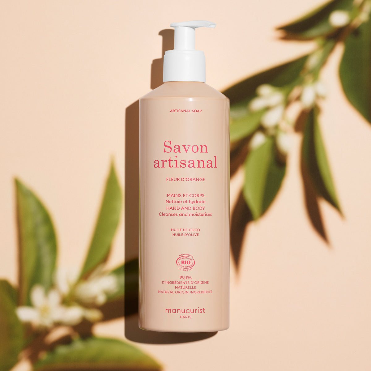 Un flacon-pompe beige de gel lavant mains et corps Manucurist à la fleur d’oranger bio - Cosmos Organic se tient droit sur un fond pêche, entouré de feuilles vertes et de leurs ombres, mettant en valeur ce savon artisanal infusé à la fleur d’oranger.