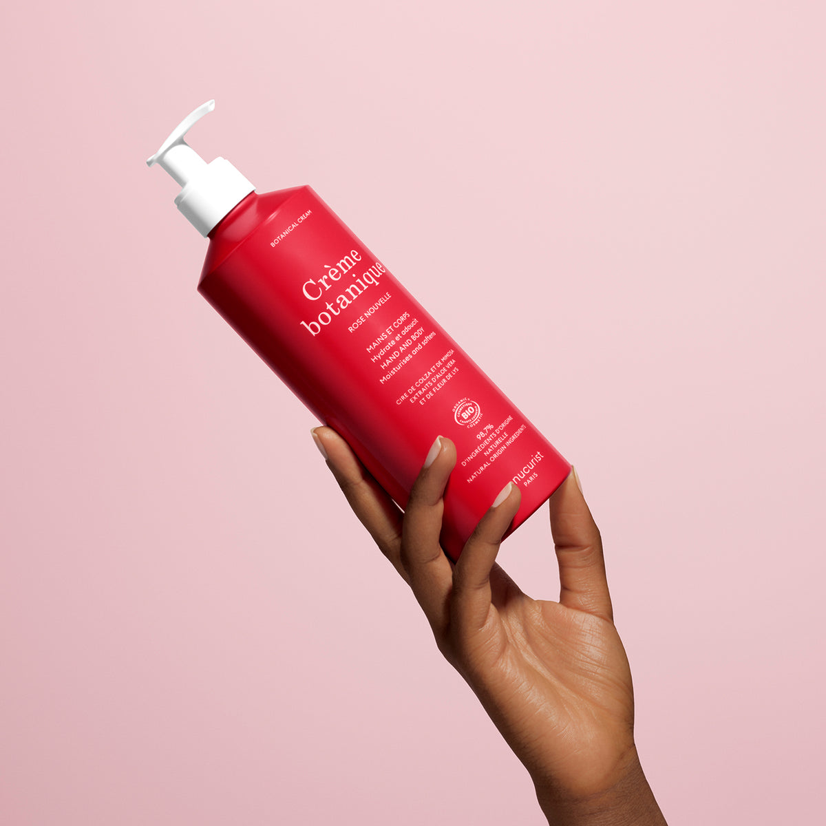 Une main à la peau foncée tient une Soothing Rose Hand Body Cream - Cosmos Organic Manucurist rouge vif avec une pompe blanche, sur un fond rose pâle. L’étiquette présente un texte blanc et le produit est conçu pour hydrater intensément la peau.