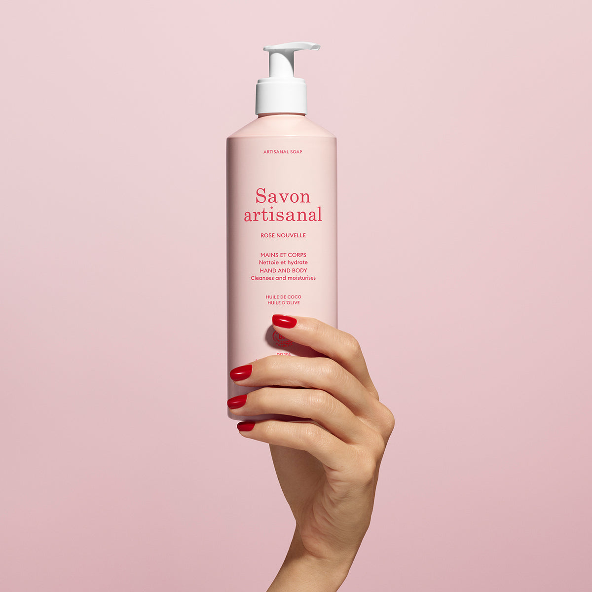 Une main aux ongles vernis de rouge tient un flacon rose de Manucurist Organic Rose Hand Body Wash - Cosmos Organic, avec une pompe blanche et du texte rouge, sur un fond rose pâle.
