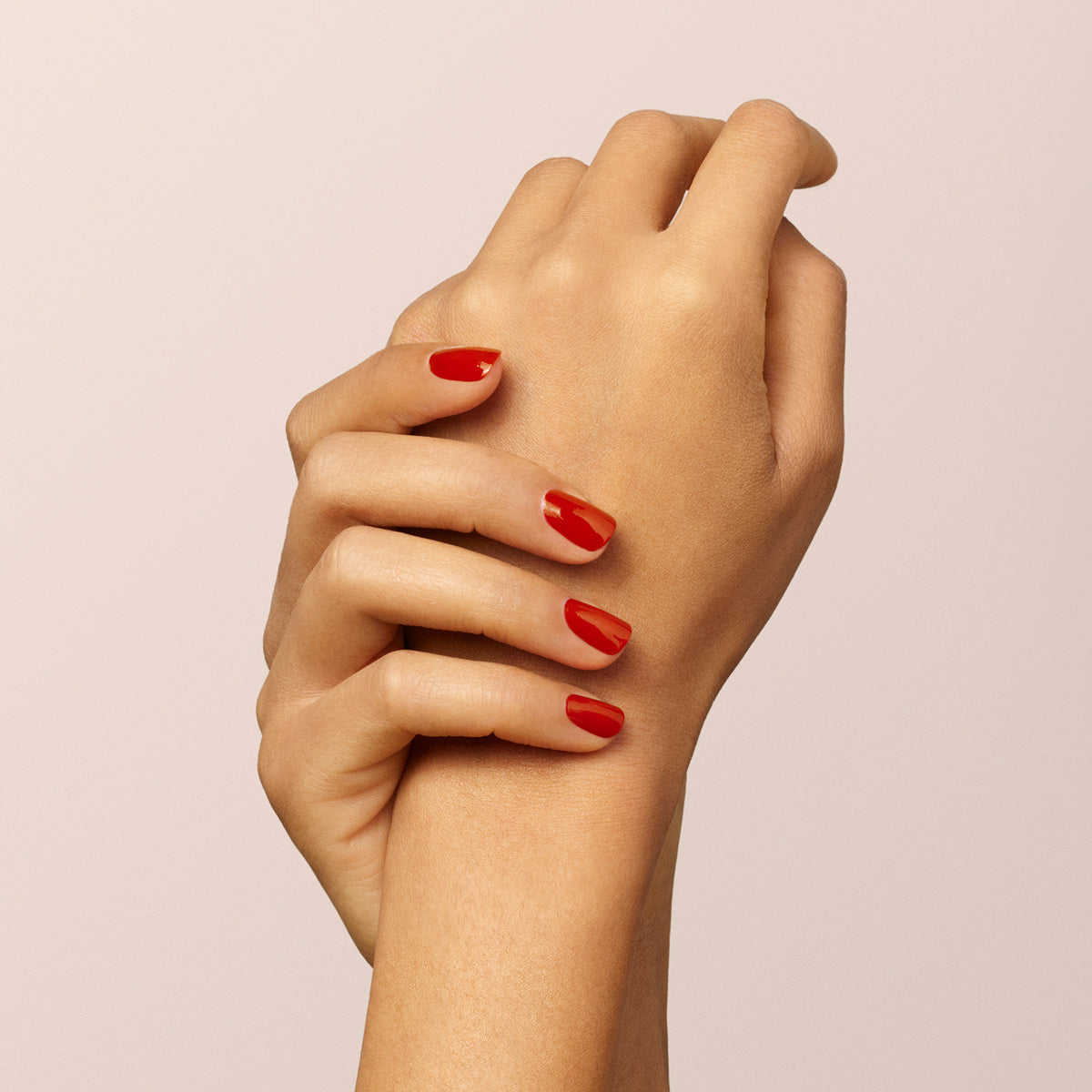 Deux mains sont présentées sur un fond clair uni. L’une tient délicatement le poignet de l’autre, mettant en valeur des ongles soigneusement manucurés peints avec Indian Summer de Manucurist, un vernis gel rouge brique brillant.