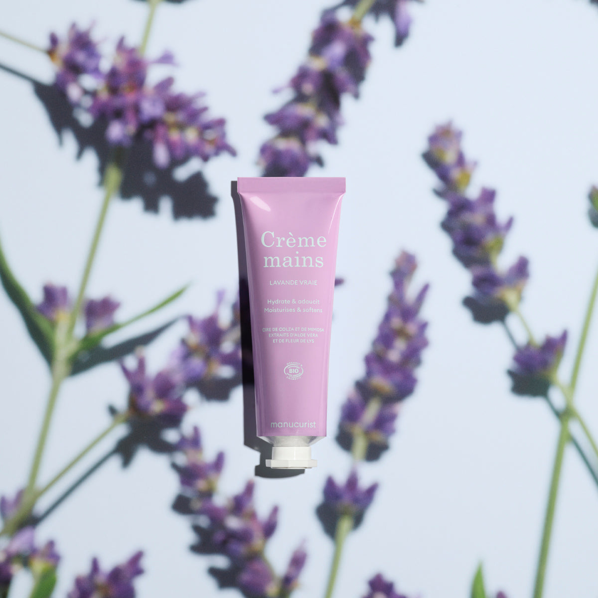 Un tube rose de Crème Mains Lavande Quotidienne Manucurist - Cosmos Organic est centré sur un fond clair avec des brins de lavande, projetant de douces ombres. Cette crème mains bio offre un parfum de lavande frais et apaisant.