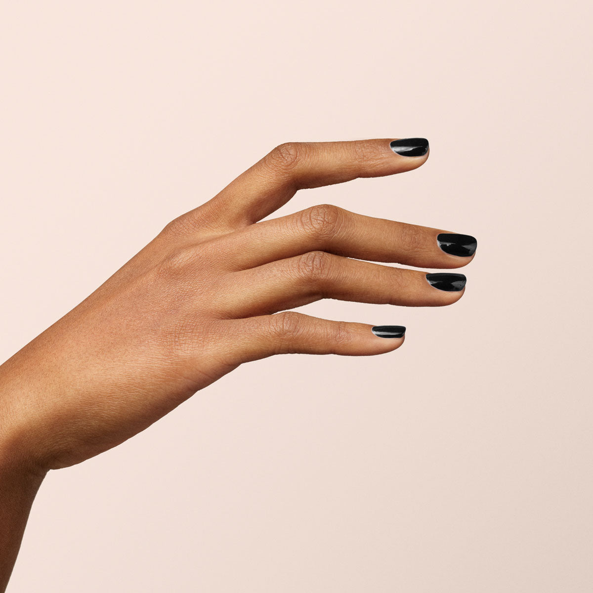 Une main à la peau mate est présentée sur un fond beige clair. Les ongles courts et soignés sont vernis avec Licorice de Manucurist pour une manucure noire chic et élégante.