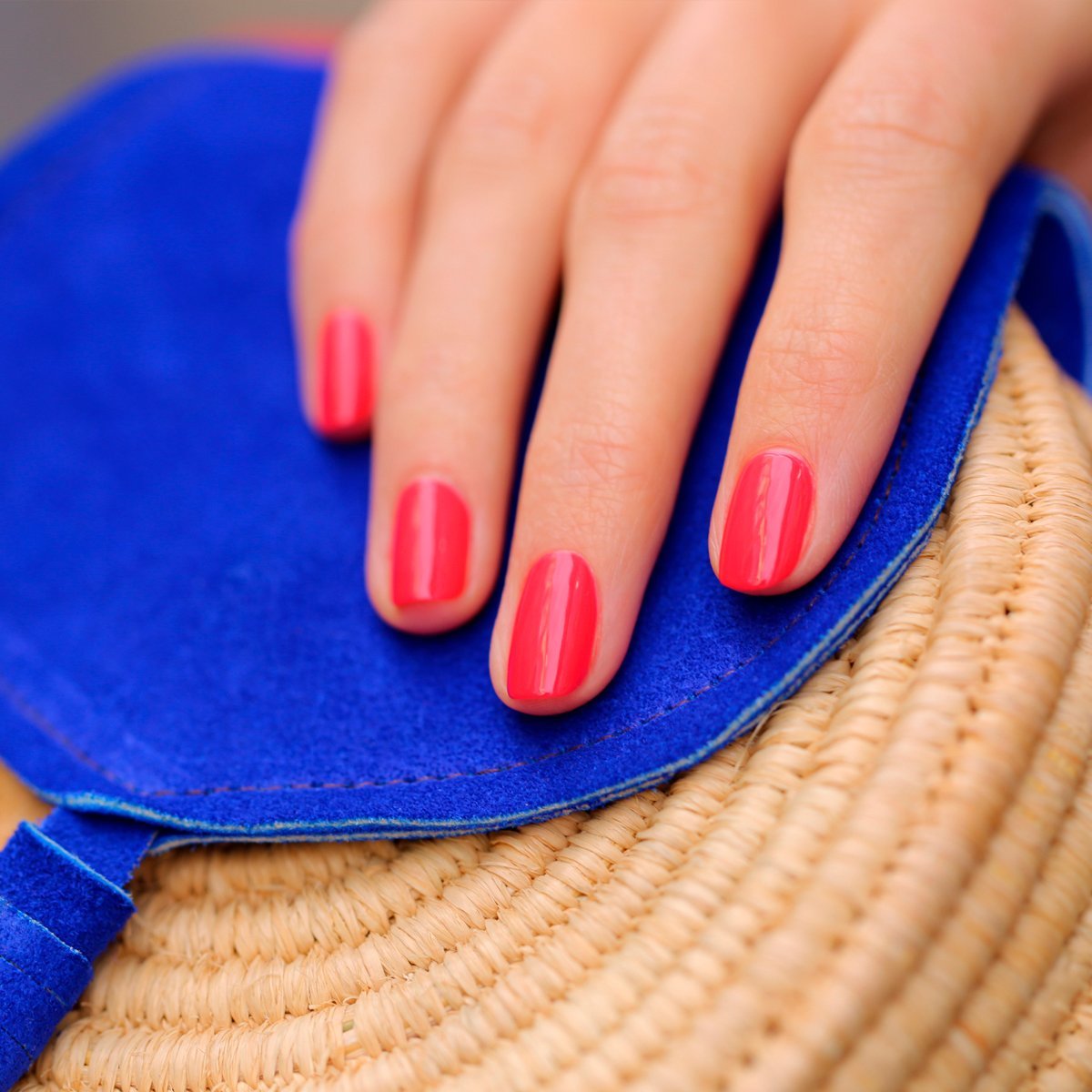 A hand with neatly manicured nails painted in Manucurist’s Bougainvillea, a vivid reddish pink polish, rests on a blue and straw-colored woven bag.