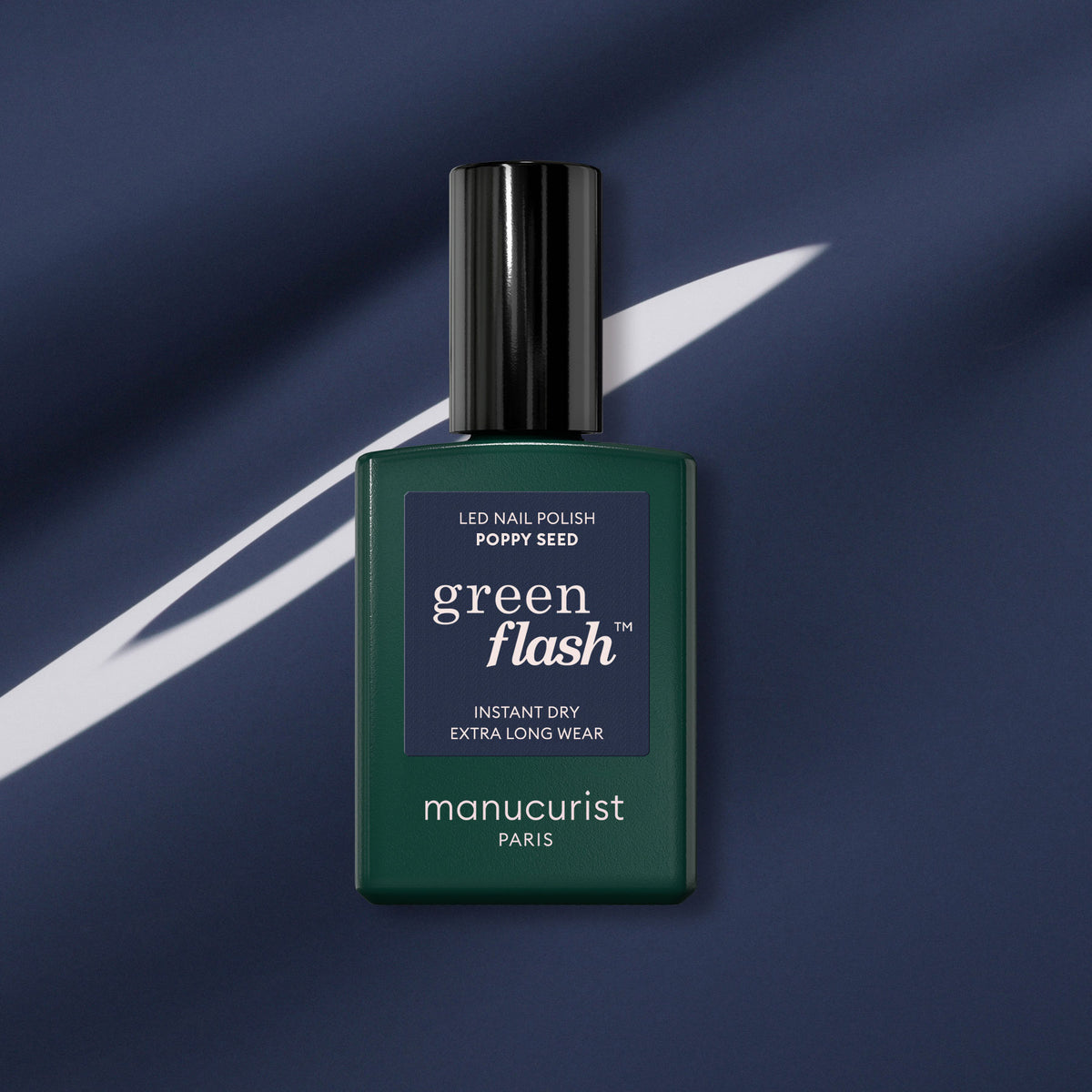 Un flacon vert foncé de vernis gel Manucurist Poppy Seed est présenté sur un fond de tissu bleu foncé avec une strie diagonale blanche.