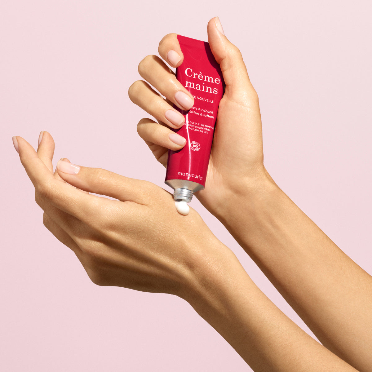 Une personne presse la Crème mains Manucurist Duo Rose Hand Cream Rose Balm - Cosmos Organic d’un tube rouge étiqueté Crème mains sur sa main, sur un fond rose clair. Cette crème aide à garder la peau douce et hydratée.