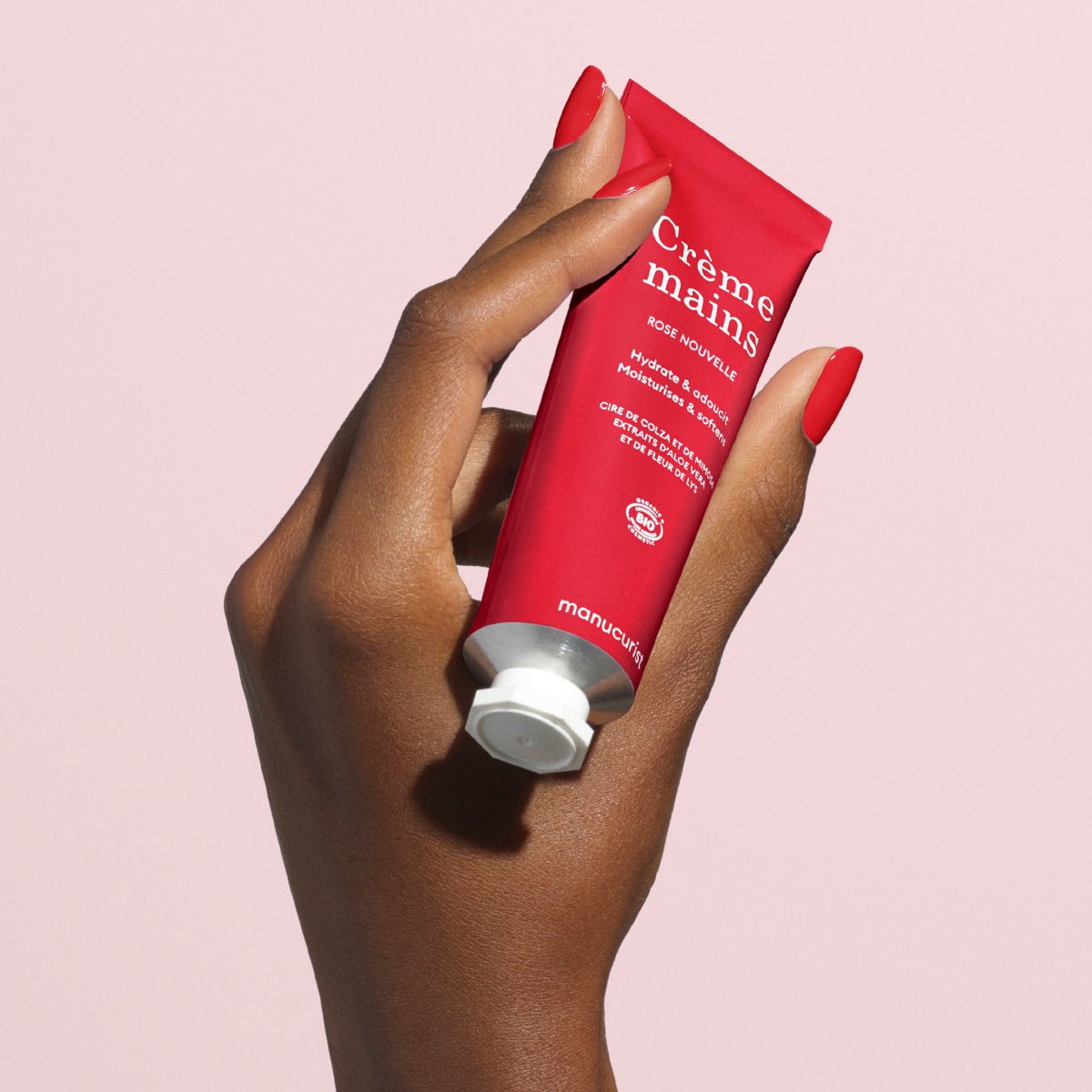 Une main aux ongles rouges tient un tube rouge de crème pour les mains Manucurist Everyday Rose - Cosmos Organic, avec un texte blanc et un bouchon blanc, sur un fond rose clair. La crème hydratante a un subtil parfum de rose.