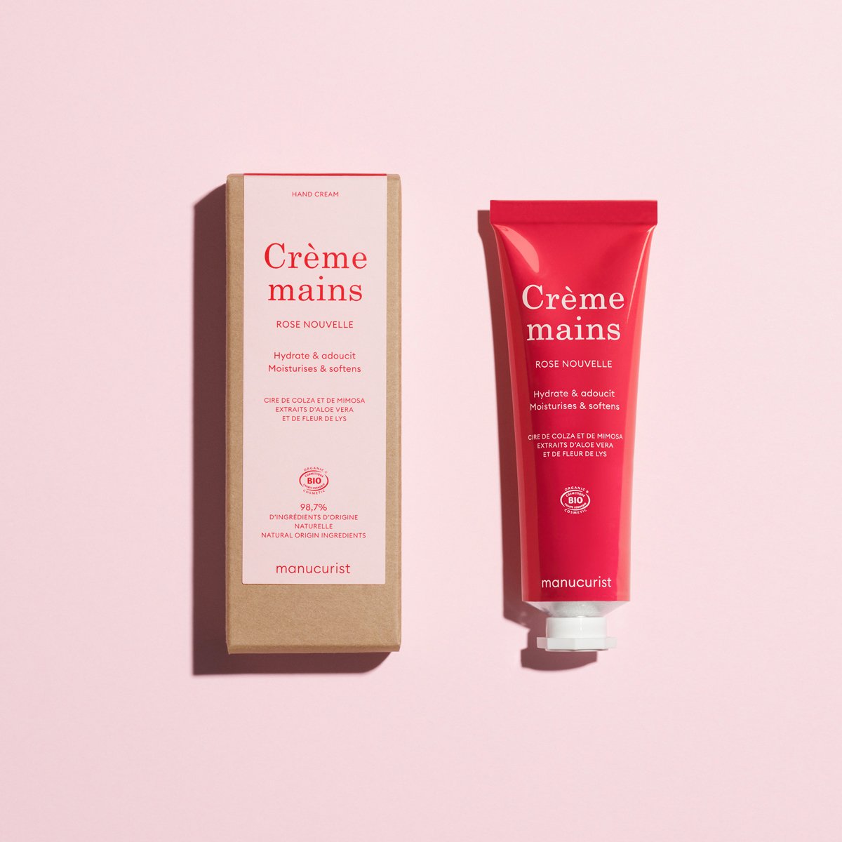 A red tube of Manucurist Everyday (Rose) Hand Cream - Cosmos Organic and its box sit on a pale pink surface. The packaging emphasizes hydration, softness, and a delicate rose scent from natural ingredients.