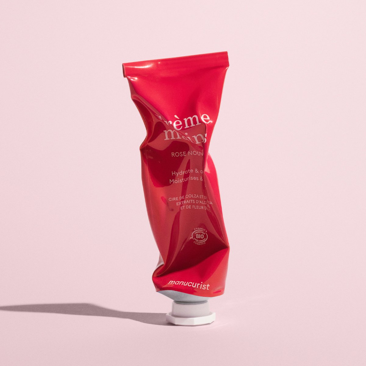Un tube rouge froissé de crème pour les mains Everyday Rose de Manucurist - Cosmos Organic, avec un bouchon blanc, se tient debout sur un fond rose. Le tube affiche « crème mains », « Hydrate Moisturise » et d’autres détails du produit.